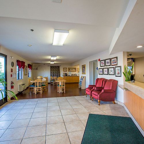 This image shows a lobby area with a reception desk, seating, plants, and a dining area in the background, featuring tiled and wooden flooring.