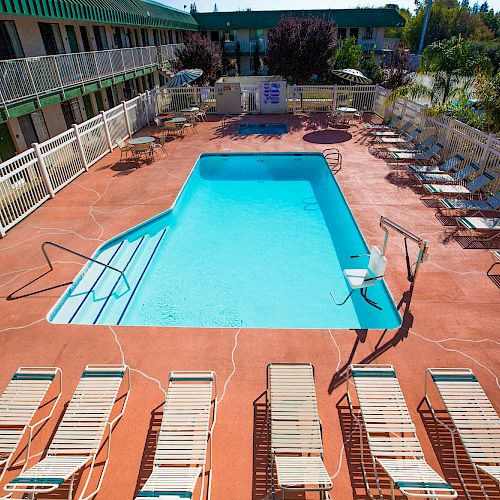 An outdoor pool area with lounge chairs, fencing, and a second-floor walkway. The area is clean and well-maintained, with a bright atmosphere.