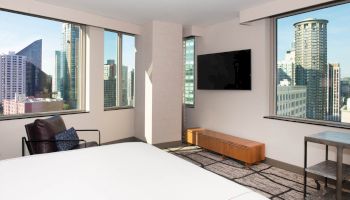 A modern hotel room with large windows showcasing a cityscape view, equipped with a bed, armchair, wall-mounted TV, and wooden console.