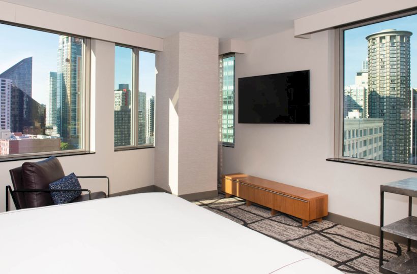 A modern hotel room with large windows showcasing a cityscape view, equipped with a bed, armchair, wall-mounted TV, and wooden console.
