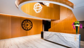 Modern reception area with a marble desk, wooden walls, large clock, spiral staircase, and decorative hanging lights.