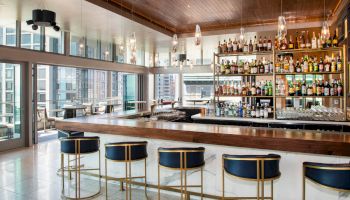 The image shows a modern bar with a sleek counter, stools, and a well-stocked back shelf. Large windows provide a city view in the background.