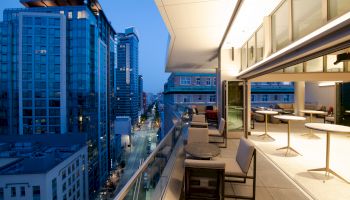 A modern cityscape at dusk, viewed from a building's terrace with outdoor seating and tables, overlooking busy streets and tall buildings.