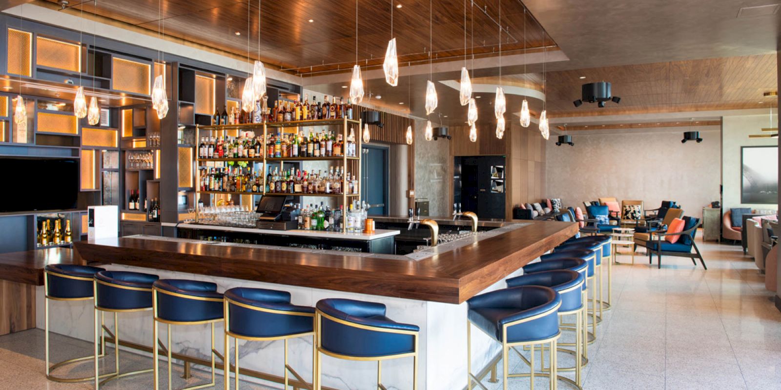 A modern bar with blue stools, a wooden countertop, and pendant lights. The shelves are stocked with various bottles, and seating areas are visible in the background.