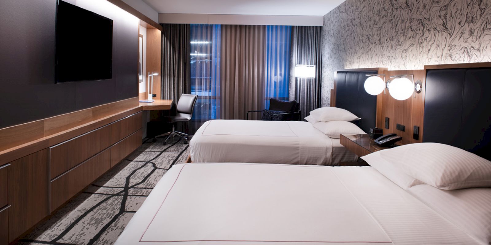 A modern hotel room features two beds, a flat-screen TV, a desk with a chair, and bedside lamps, with an overall neutral and stylish decor.