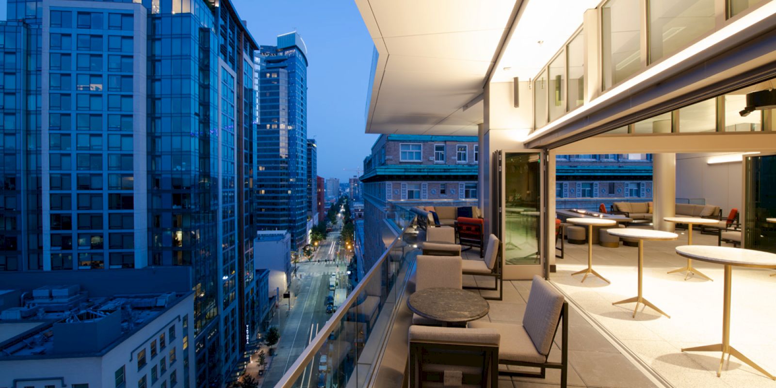 A modern balcony with tables and chairs, overlooking a street flanked by tall buildings during twilight, city lights starting to illuminate.