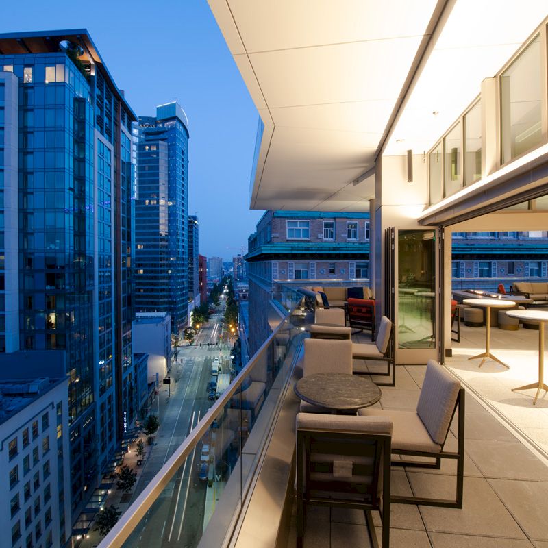 A modern cityscape at dusk, featuring high-rise buildings and a furnished balcony area with tables and chairs, offering a scenic urban view.