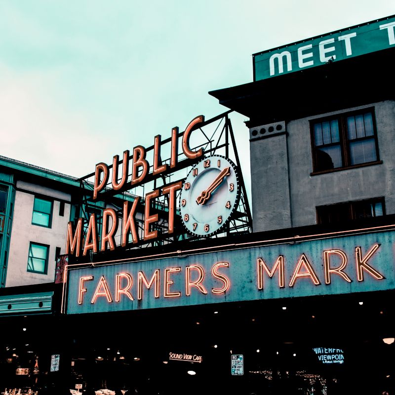 A vibrant photo of a public market with a neon "FARMERS MARKET" sign and a clock, surrounded by older buildings in an urban setting.