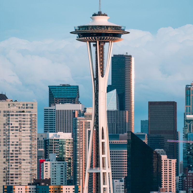 The image shows the Space Needle, a prominent observation tower, with the surrounding skyline of Seattle, including various skyscrapers, under a cloudy sky.