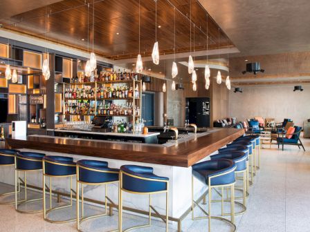A modern bar with a wooden countertop, blue bar stools, and hanging lights. Shelves of liquor are in the background with a seating area to the right.