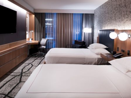 The image shows a modern hotel room with two beds, a desk, a TV, and contemporary decor with wall lamps and patterned wallpaper.