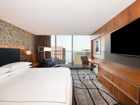 A modern hotel room with a large bed, wall-mounted TV, desk, and chair. A window overlooks city buildings and a body of water.