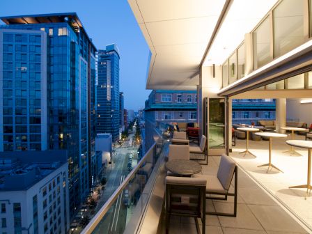 This image shows a modern balcony overlooking an urban street at dusk, with seating and tables arranged outside an open indoor area.