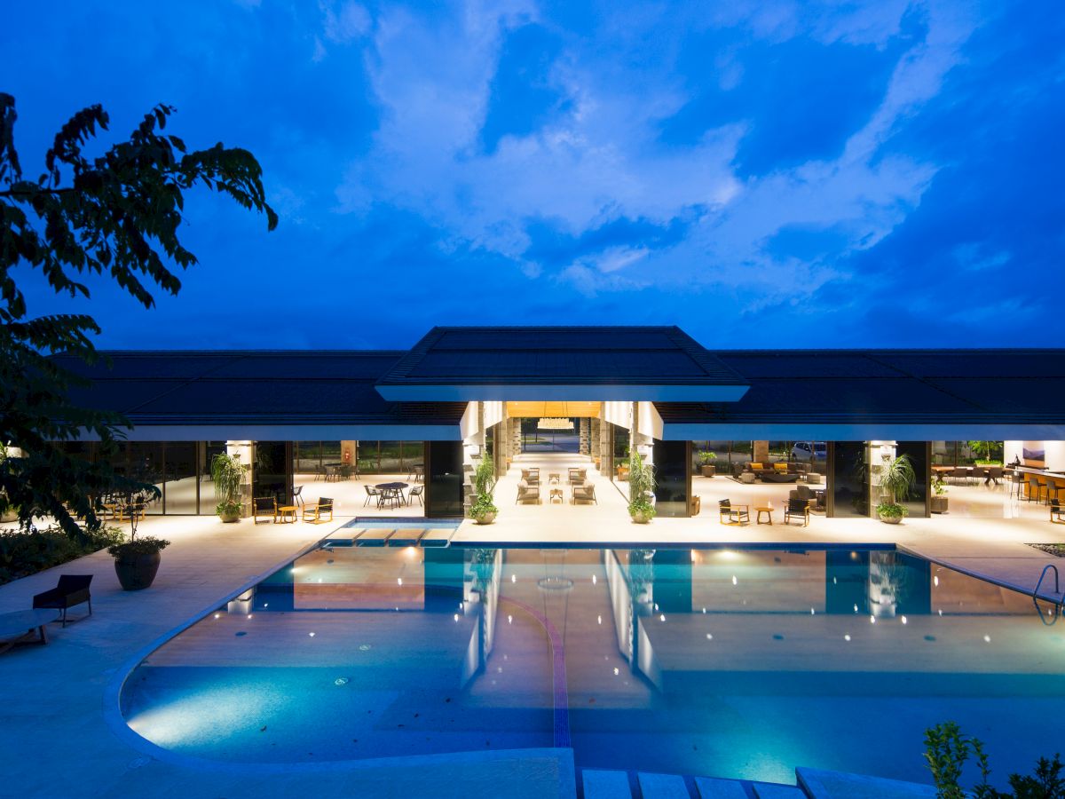 A luxurious resort building with lights on, featuring a large, illuminated swimming pool in the foreground, set against a twilight sky.