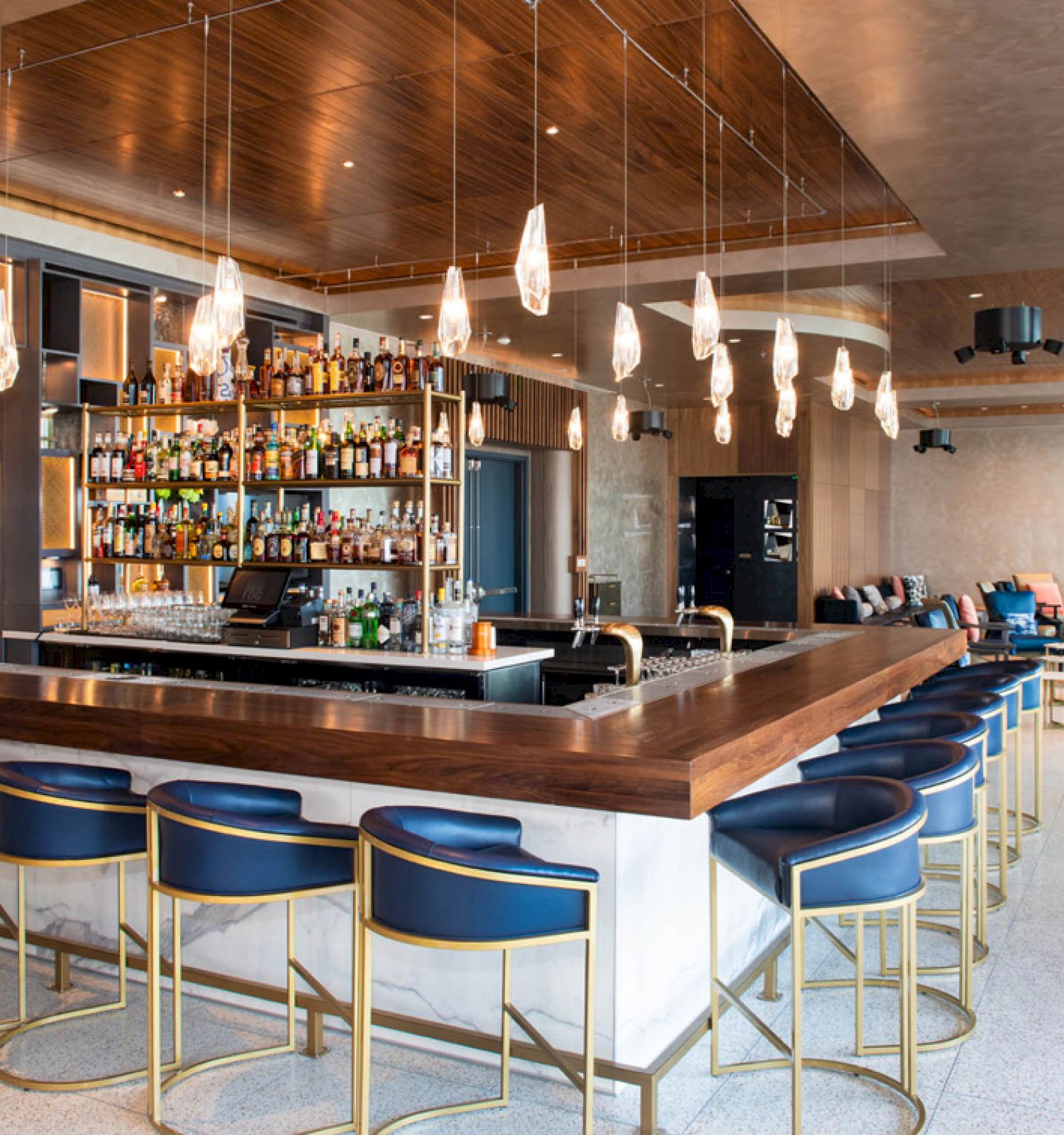 The image features a modern bar with blue cushioned stools, a well-stocked bar, hanging lights, and a lounge area with seating in the background.