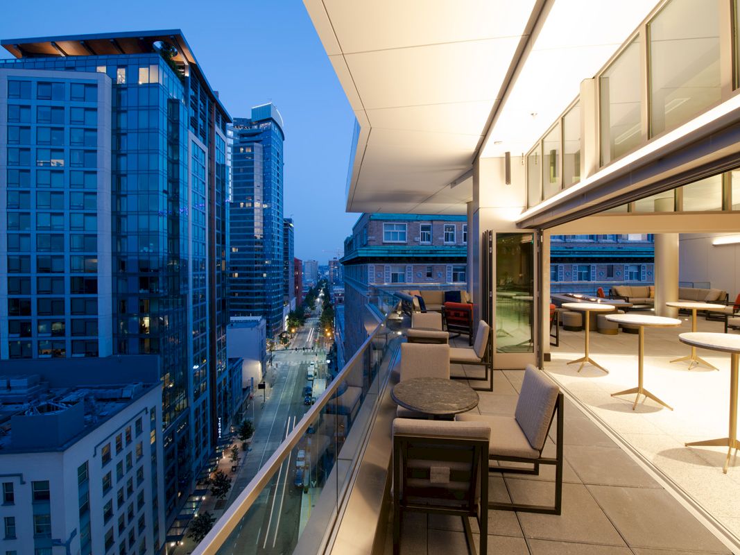 A modern, elevated city view featuring a balcony with seating, overlooking a street lined with tall buildings during twilight.