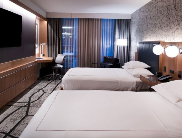 The image shows a modern hotel room with two neatly made beds, a flat-screen TV, a desk, and large windows with curtains.