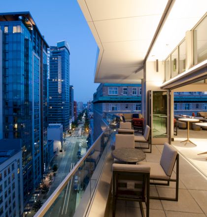 A modern cityscape featuring tall buildings and a furnished balcony with tables and chairs overlooking the street below at dusk.