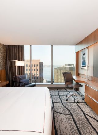 A modern hotel room featuring a large bed, wall-mounted TV, desk, chair, and a window with a city view.