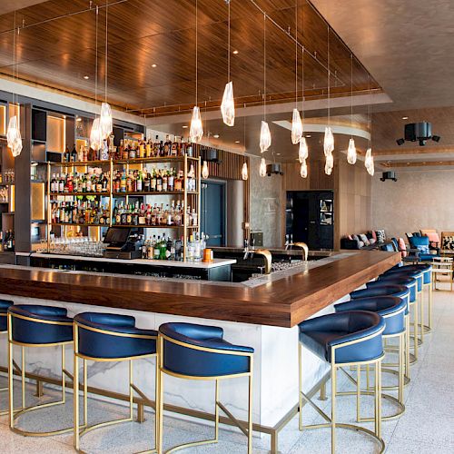 The image shows a modern bar with a wooden countertop, blue chairs with gold accents, hanging lights, and a stocked back bar.