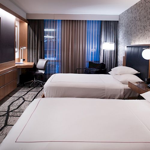 The image shows a modern hotel room with two double beds, a flat-screen TV, a desk with a chair, and a wall with decorative wallpaper.