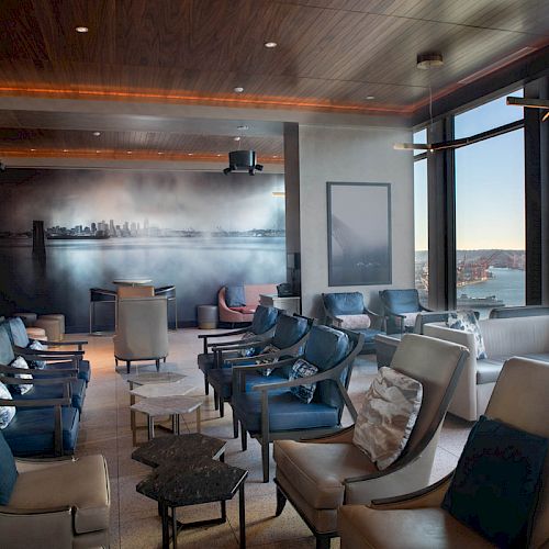 A modern lounge with plush seating, large windows offering a waterfront view, and wall art depicting a city skyline.