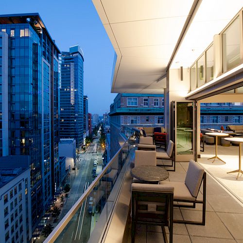 The image shows a modern cityscape view from a high-rise balcony with tables and chairs, overlooking a street lined with other buildings.