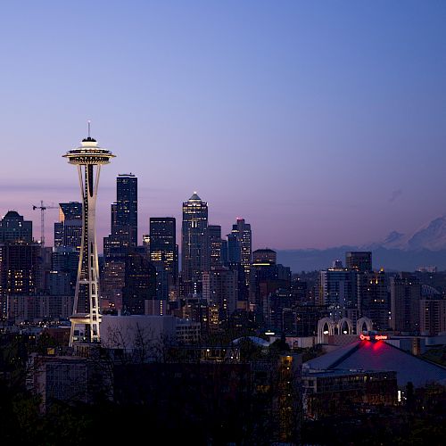 The image depicts the skyline of Seattle at dusk, highlighting the Space Needle and Mount Rainier in the distance against a colorful sky.