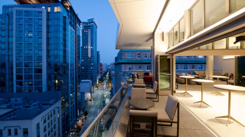 A modern balcony with tables and chairs overlooks a cityscape with tall buildings and lit streets during dusk.