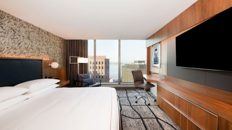 This image shows a modern hotel room with a large bed, a wall-mounted TV, a desk with a chair, and a window with a city view in the background.