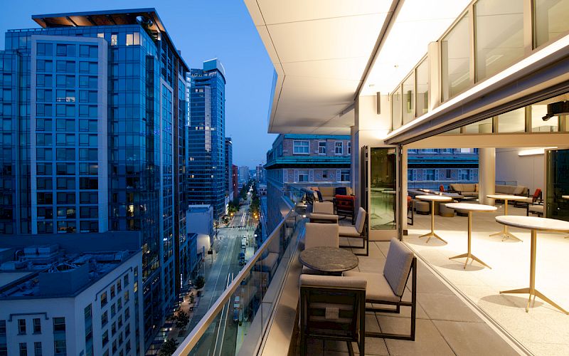 A modern rooftop terrace with seating areas overlooks a city street lined with tall buildings at dusk, with lights illuminating the scene.