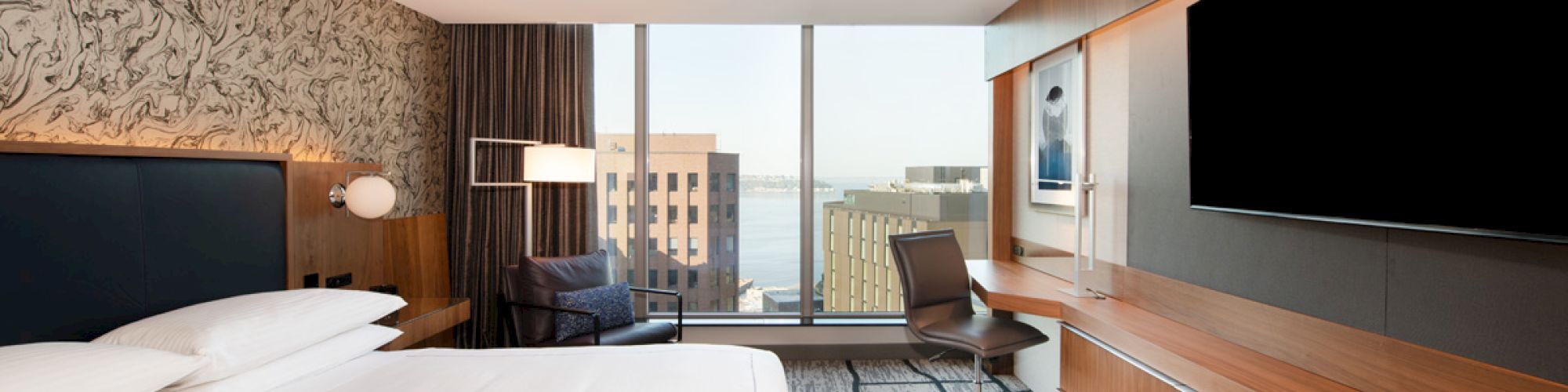 A modern hotel room with a large bed, TV, desk, chair, and floor-to-ceiling window offering a city and water view.