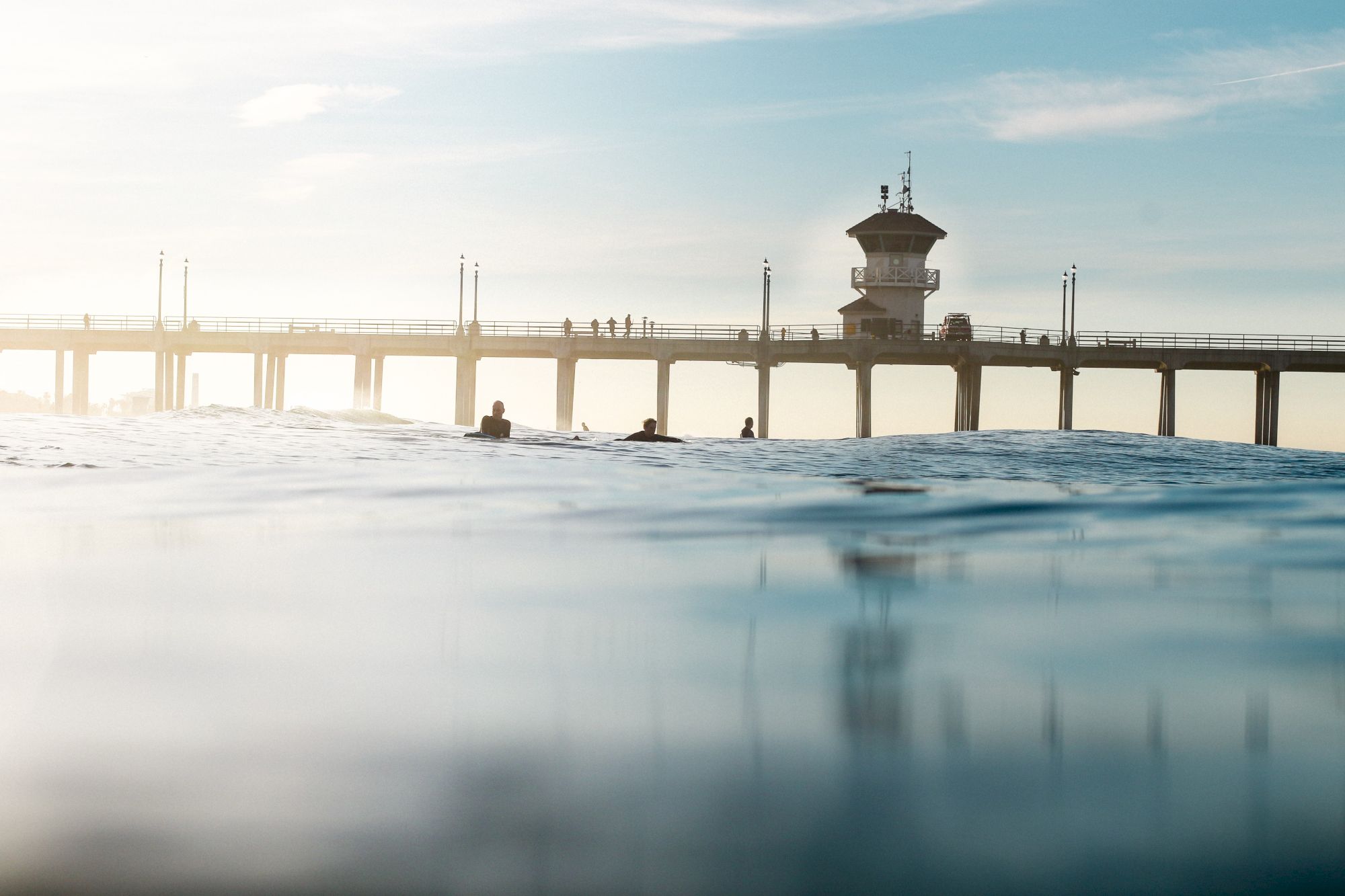 A serene image of a pier extending over the water with a few people walking on it and three individuals in the water below it, under a clear sky.